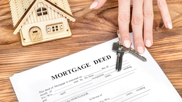A hand holding house keys above a mortgage deed with a miniature house model on a wooden desk, symbolizing a grant deed transfer.