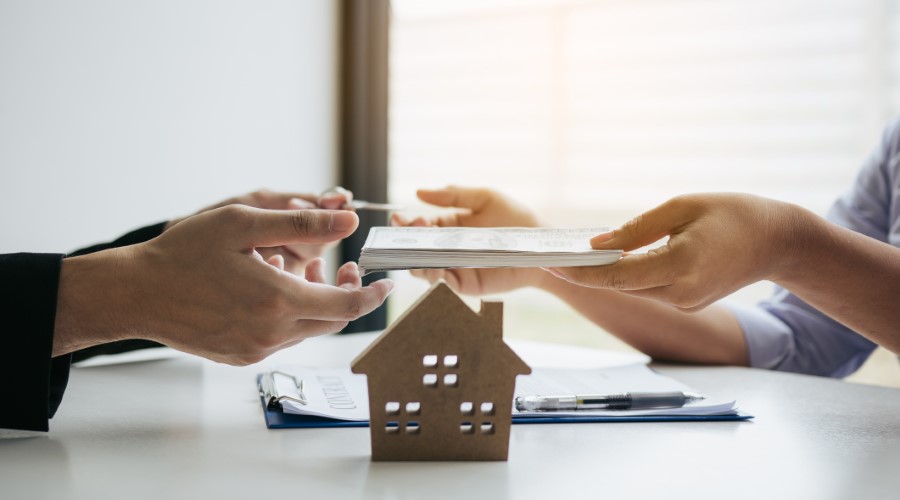 Hands exchanging a real estate document and cash over a desk with a house model, symbolizing a property transaction.